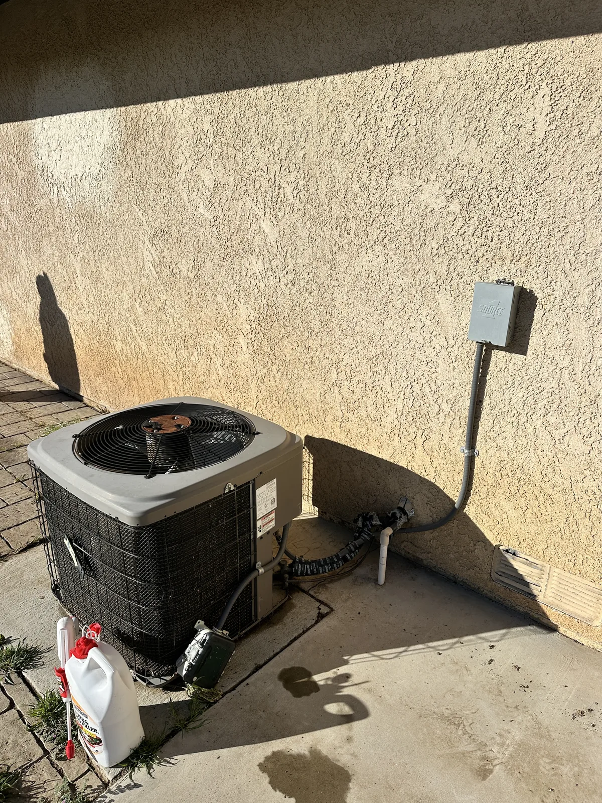 Outdoor air conditioning condenser unit on a concrete pad beside a stucco wall with an electrical disconnect box and whip in an Inland Empire backyard.