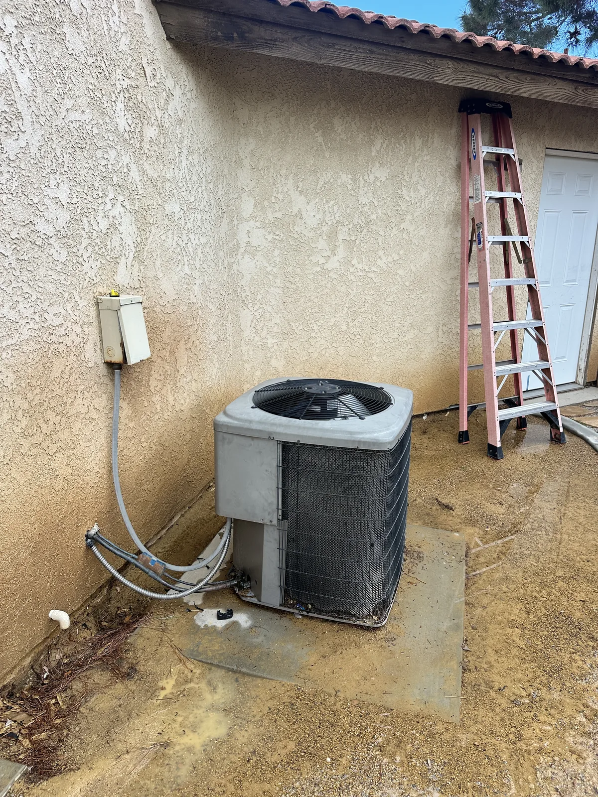 Wall-mounted air conditioning condenser unit attached to a stucco home exterior in the High Desert with refrigerant line set and a disconnect box.
