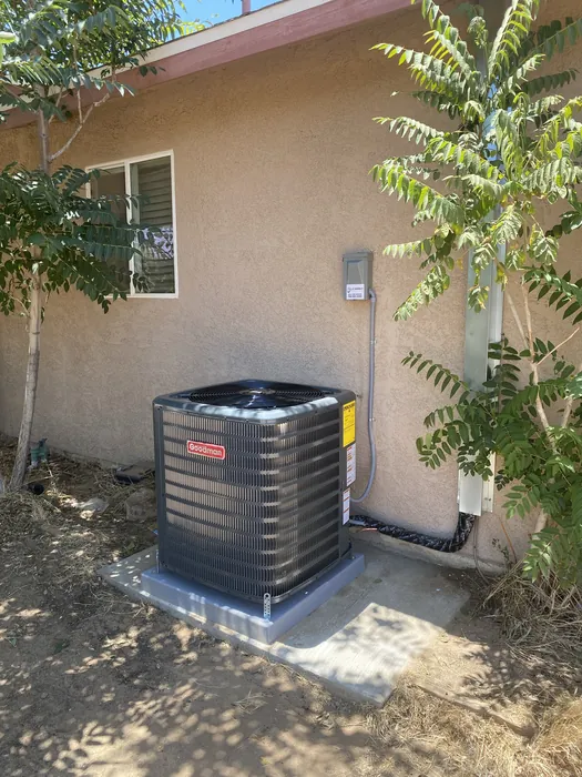 New Goodman AC condenser installed on a pad against a stucco wall with disconnect and refrigerant line set in a High Desert side yard.
