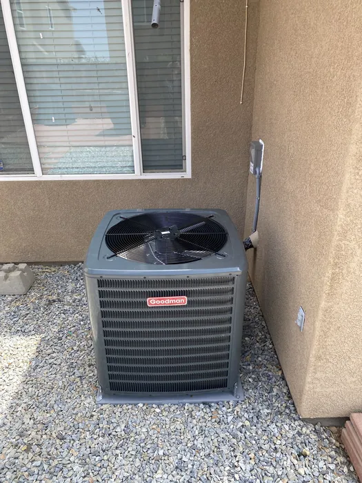 An outdoor air conditioning condenser unit with a visible Goodman brand label sits on gravel beside a stucco wall and a window.