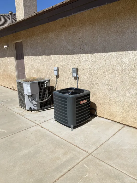 New Goodman AC condenser installed next to an older existing condensing unit on a concrete pad outside a High Desert home.