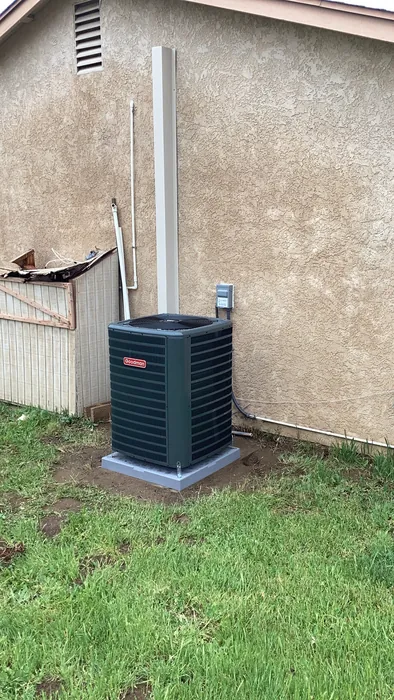 Goodman outdoor AC condenser unit installed on a pad at the edge of a backyard patio next to the grass at a High Desert home.