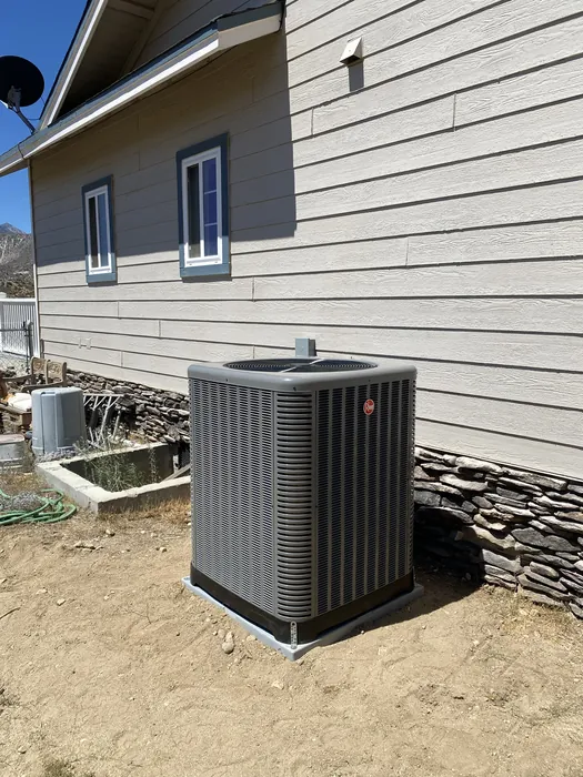 An outdoor air conditioning condenser unit installed on a gravel pad next to a home with horizontal lap siding and two small windows.