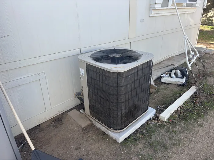 Outdoor air conditioning condenser unit set on a pad beside the exterior wall of a manufactured home with refrigerant lines and disconnect.