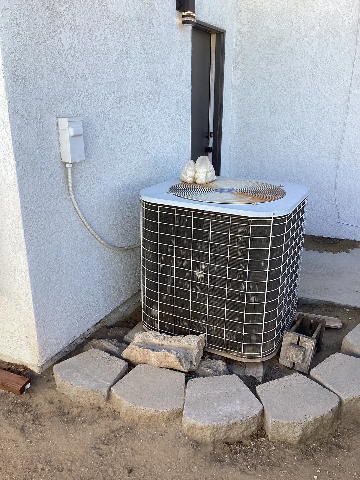 Outdoor AC condenser unit set on a stucco pad next to a stone-edged yard at a High Desert home.