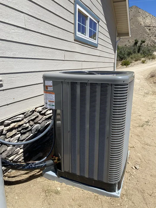 A gray packaged HVAC condenser unit sitting on a composite deck with its coil face exposed and a refrigerant line running below, hillside in the background.