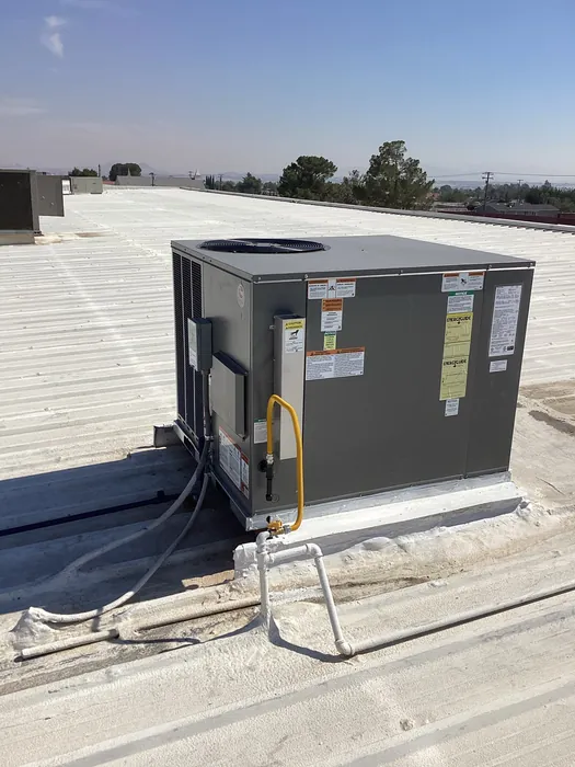 A gray packaged rooftop HVAC gas-pack unit on a concrete pad with a yellow flexible gas connector and white gas piping, set against an open sky and dirt lot.