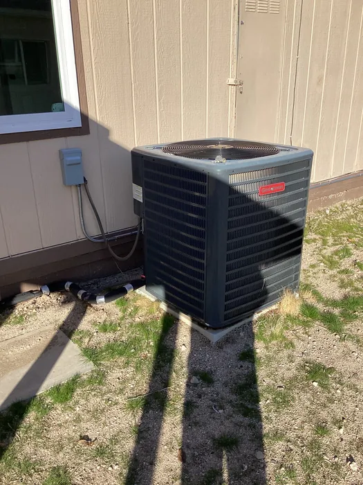 Outdoor split-system AC condenser unit installed beside a tan-sided High Desert home with a wall-mounted electrical disconnect and refrigerant line set.