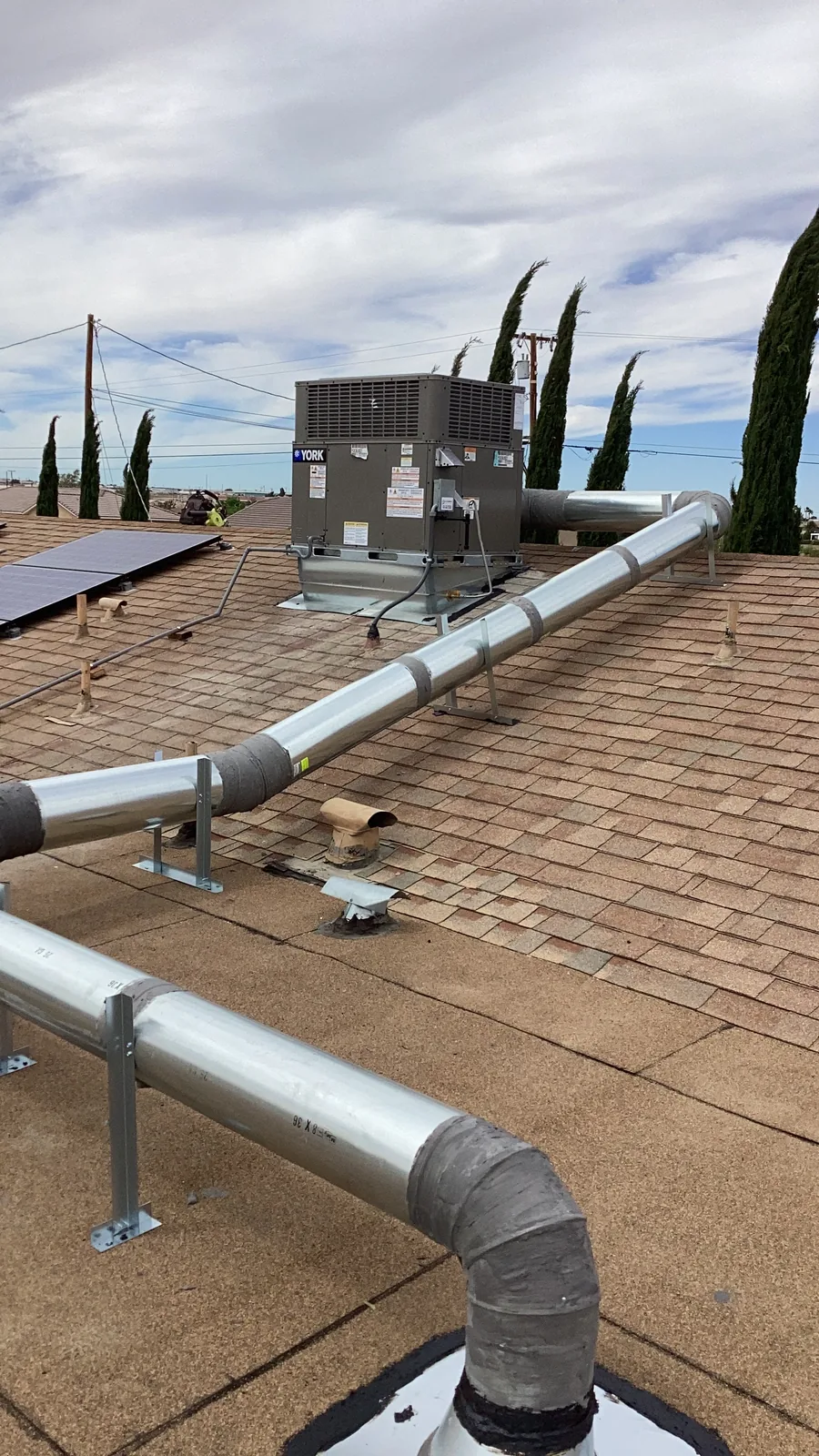Rooftop-mounted packaged AC condenser unit with freshly installed metal supply and return duct runs across composition shingles on a High Desert home.