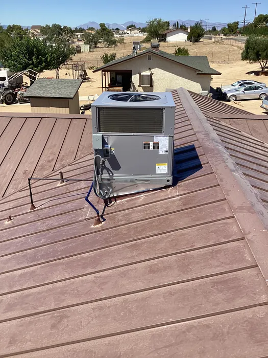 A gray packaged rooftop HVAC unit installed on a brown standing-seam metal roof of a residential home with desert neighborhood and clear sky in the background.