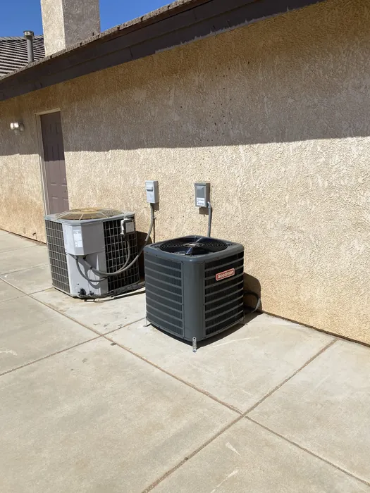 Two outdoor air conditioning condenser units sitting on a dirt pad beside a stamped concrete patio with two electrical disconnect boxes on the wall behind them