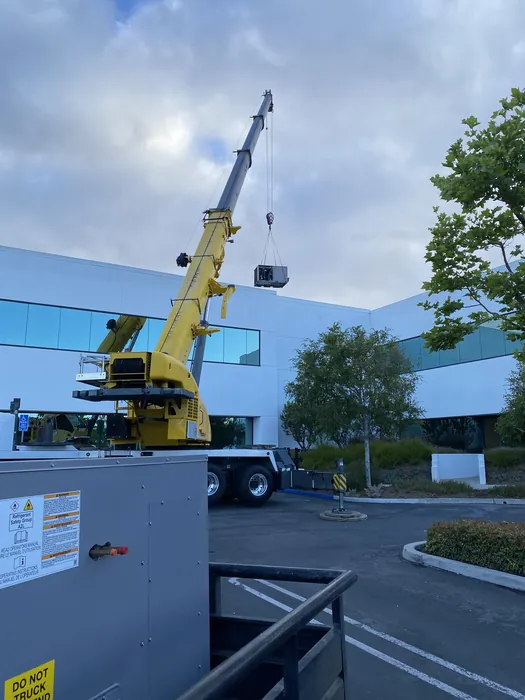 A large yellow crane lifting a commercial rooftop HVAC unit up the side of an office building during a packaged unit installation in the Inland Empire.