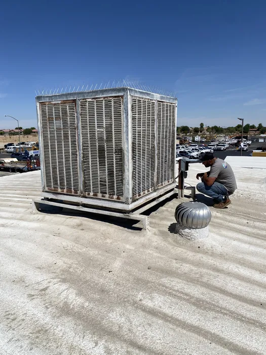 HVAC technician kneeling beside a rooftop commercial package unit and turbine vent on a flat commercial roof in the High Desert.
