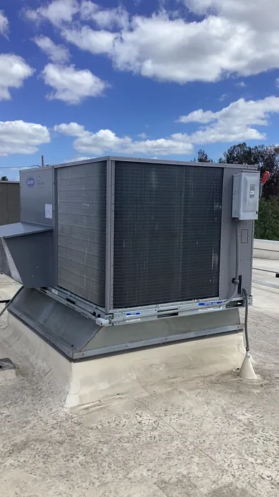 A gray commercial packaged rooftop HVAC unit with an electrical disconnect box mounted on the roof curb under a clear sky.