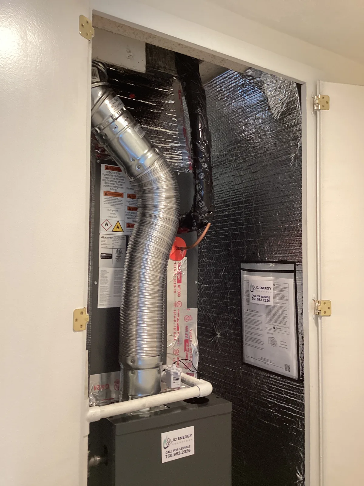 Air handler in an insulated ceiling closet with a flexible silver supply duct and white condensate lines connected to the plenum, carrying a JC Energy startup label.