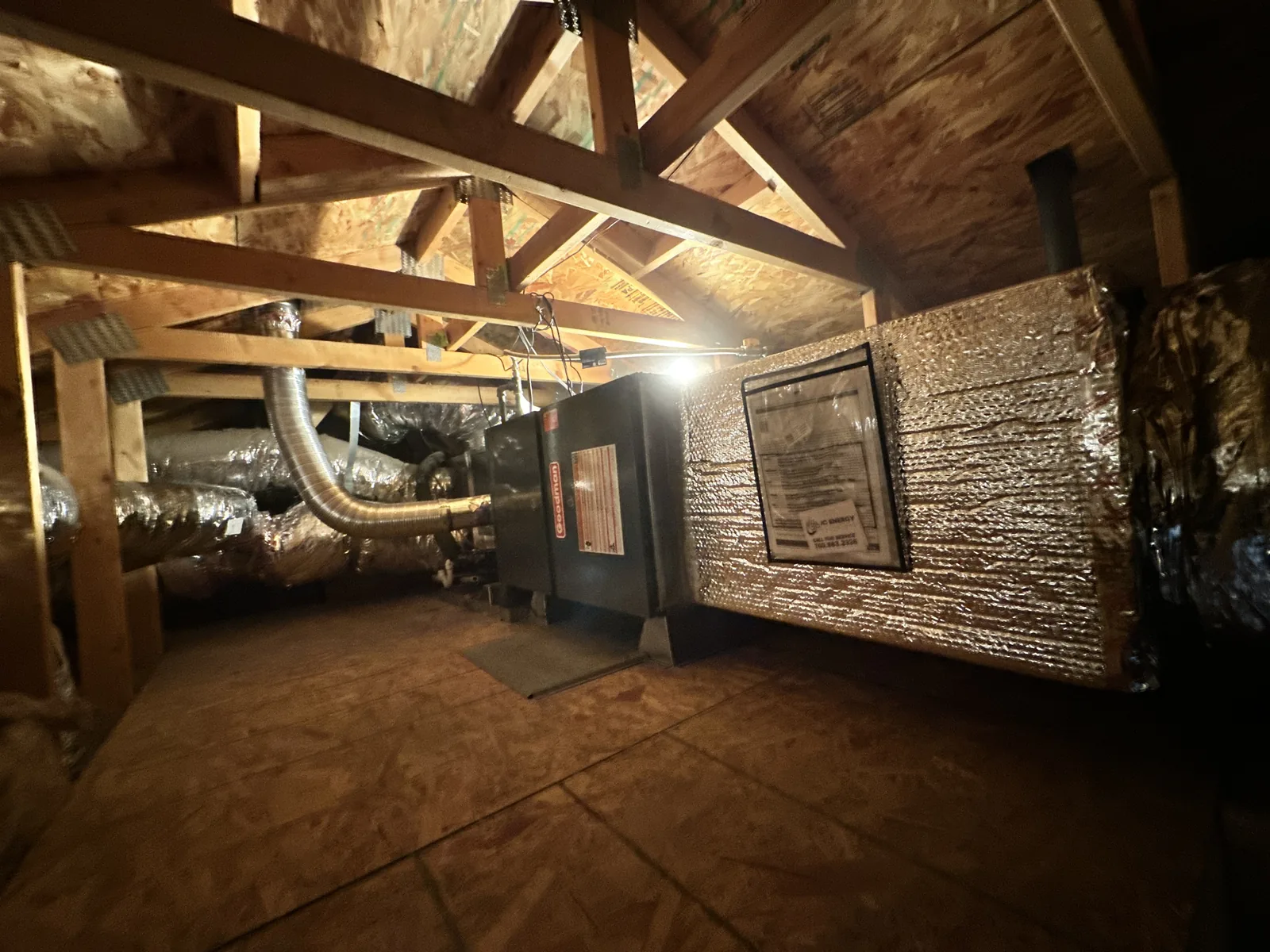 Attic furnace and air handler carrying a JC Energy service sticker, connected to insulated flex ducts and a wrapped supply plenum under wood trusses.