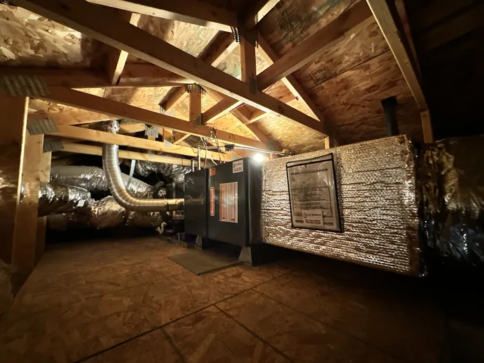 Attic-installed furnace and air handler tied into insulated flex duct and a wrapped supply plenum under exposed wood roof trusses.