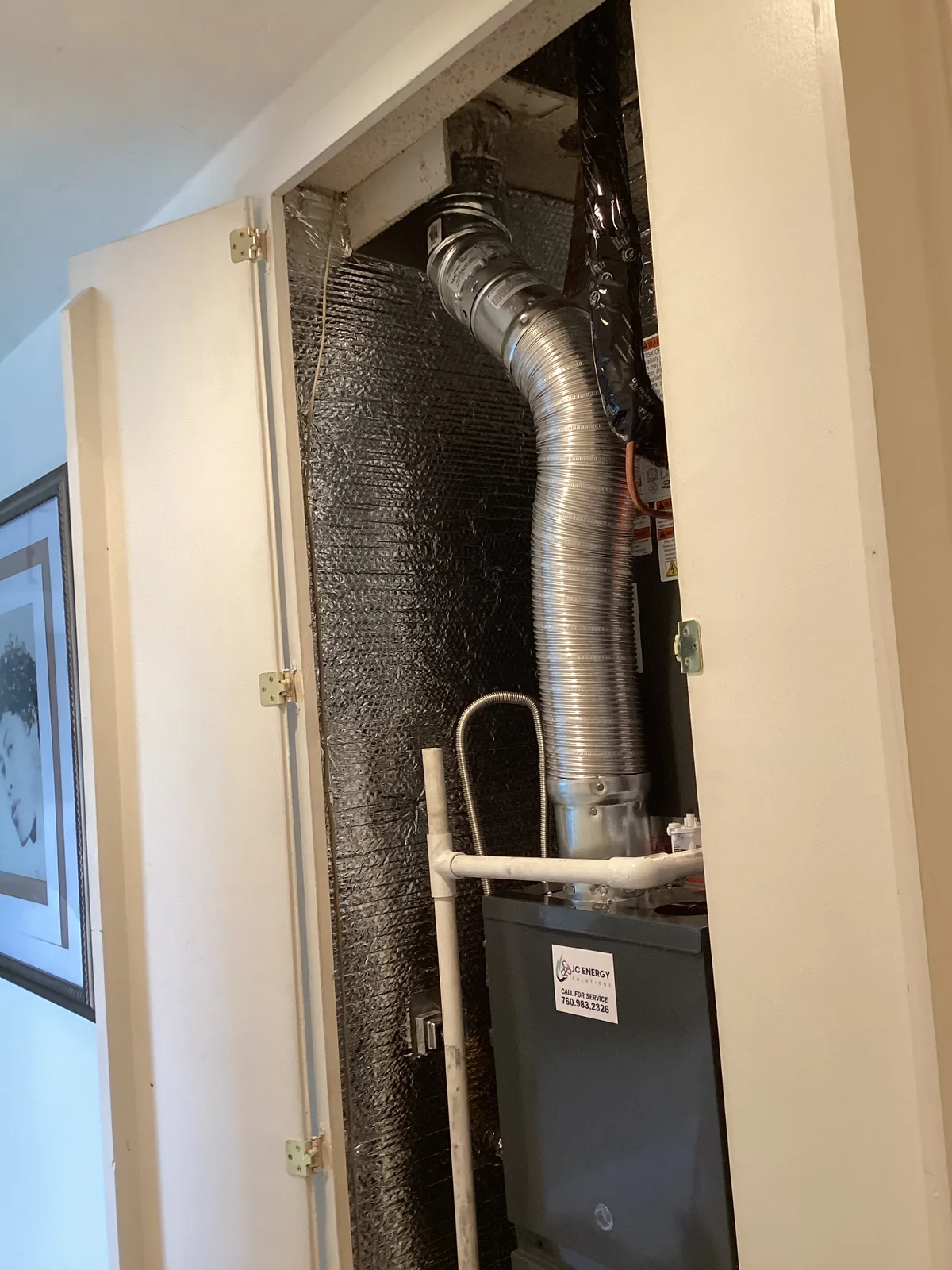 Closet-mounted air handler with a flexible metal flue duct and a JC Energy service sticker visible behind an access panel in a High Desert home.