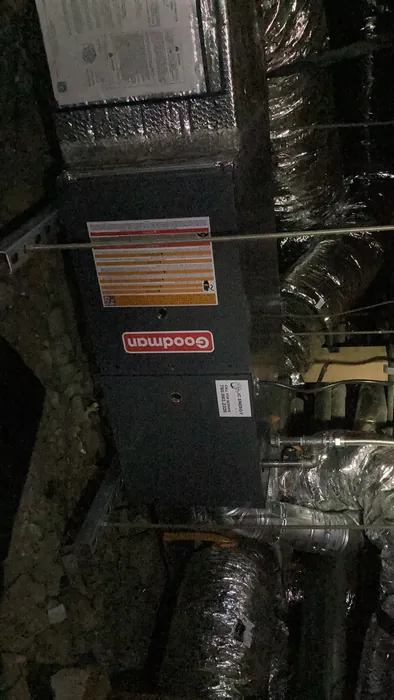 A Goodman gas furnace and air handler installed in an attic surrounded by insulated flex ductwork with a JC Energy service sticker on the cabinet.