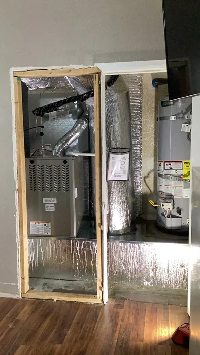 Newly installed Goodman furnace and air handler with foil-faced insulation and supply ductwork in a framed interior closet beneath a water heater.