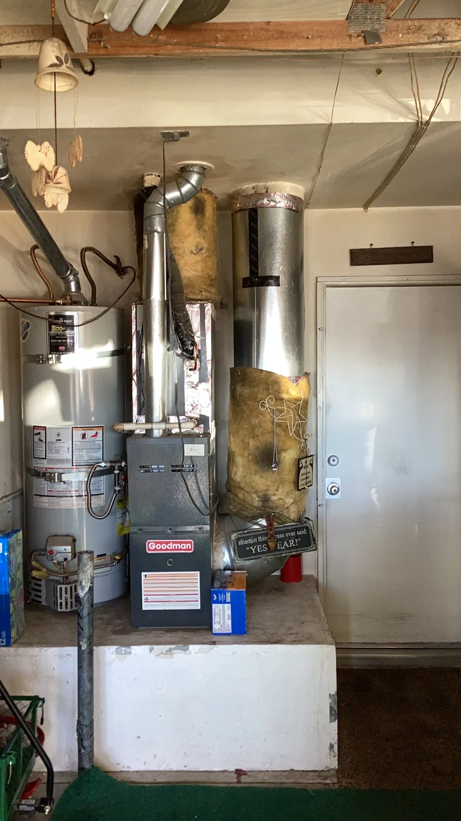 Goodman gas furnace installed beside a tank water heater in a garage with metal flue venting overhead in an Inland Empire home.