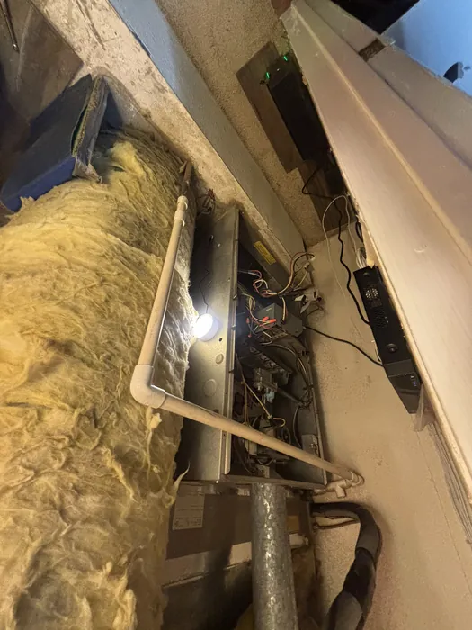 Open blower and wiring compartment of a horizontal furnace in a tight insulated crawlspace lit by a work light with PVC condensate piping overhead.