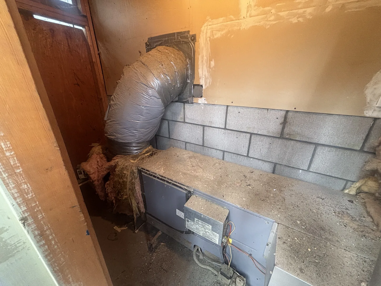 An older horizontal gas furnace with a sagging flex duct and exposed wiring against a block wall in a High Desert garage, staged for replacement.