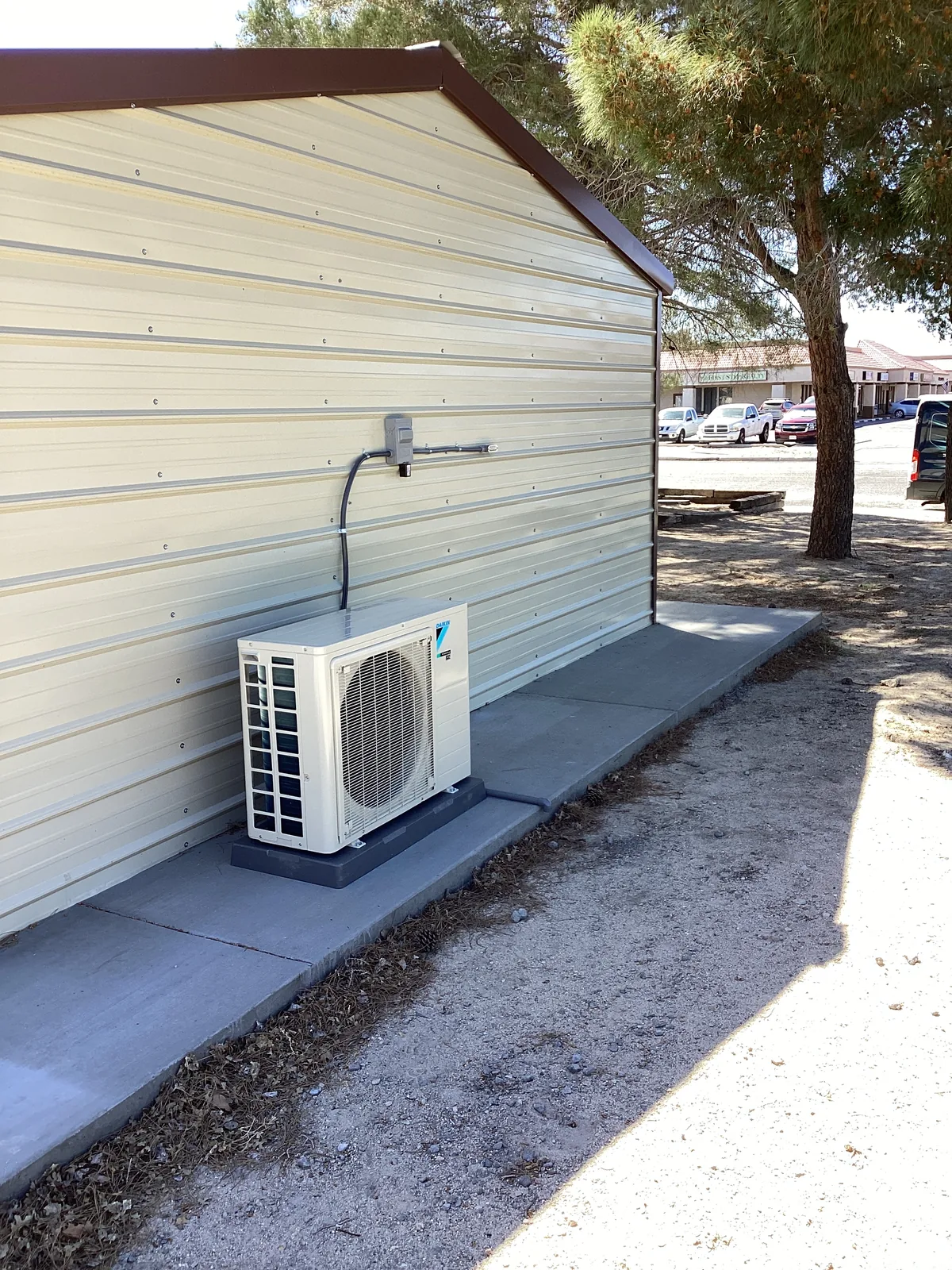 Daikin mini-split condenser mounted on a wall bracket along a metal building wall in a residential side yard with bare trees.
