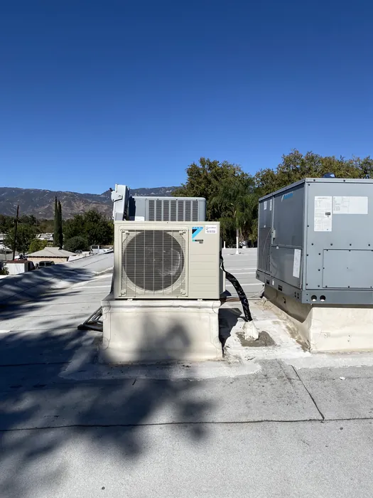 A cream-colored Daikin ductless mini-split outdoor condenser unit on a concrete pad outdoors, with a larger gray packaged HVAC unit behind it under a clear blue sky.