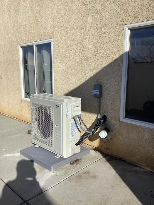 Wall mounted mini split heat pump condenser on a gray bracket with insulated lineset and disconnect box outside a stucco High Desert home
