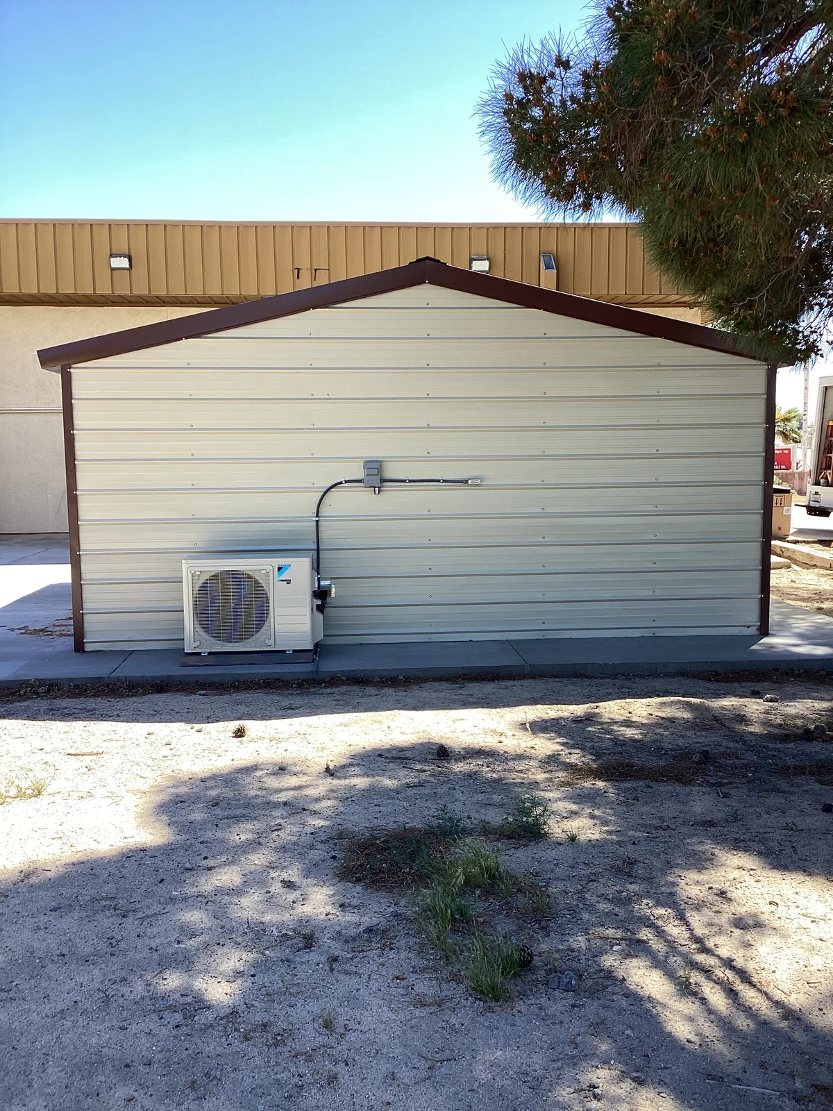 Wall-mounted ductless mini-split outdoor condenser on a metal shop building with sandy High Desert ground and pine trees.