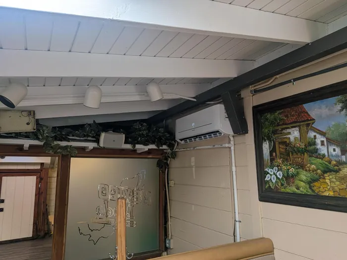 White ductless mini-split wall head mounted high on an interior wall near a painted beam and framed cottage artwork inside a restaurant dining room.