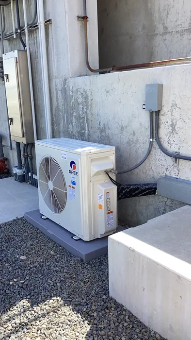 A Gree mini-split outdoor condenser unit installed on a pad against a concrete wall with electrical disconnect and refrigerant line set.