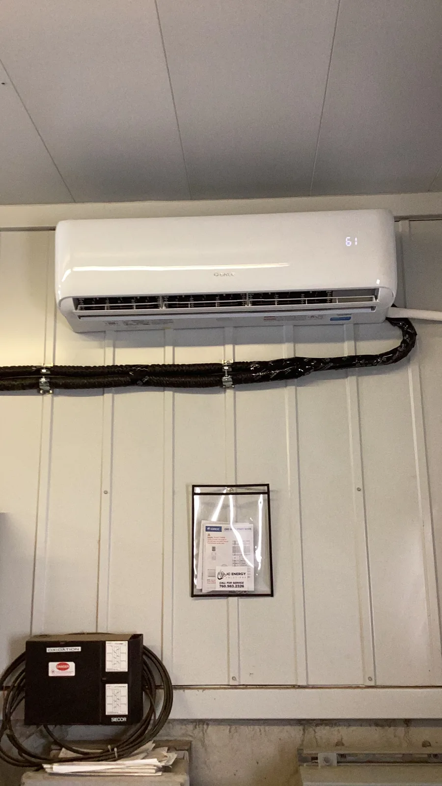 An indoor wall-mounted mini-split head with a flexible insulated line set running down to a disconnect box on a metal wall.