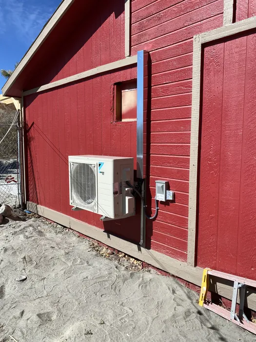 JC Energy Solutions mini split outdoor condenser mounted on the wall of a red building on sandy High Desert ground.