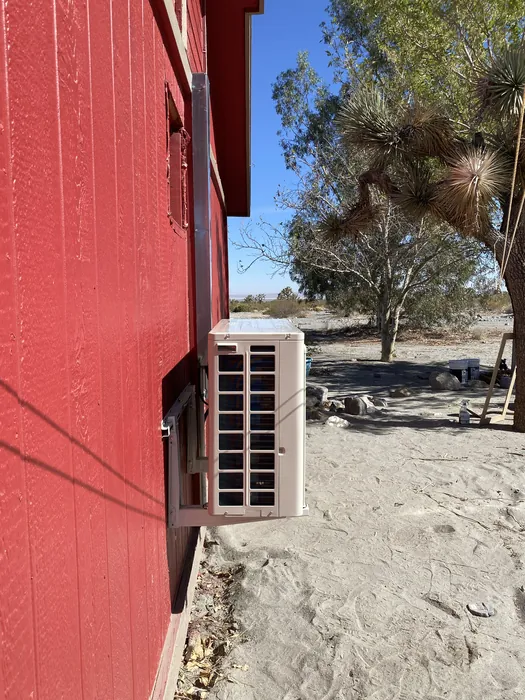 A ductless mini-split outdoor unit on red wood siding with a Joshua tree and open desert yard behind it.