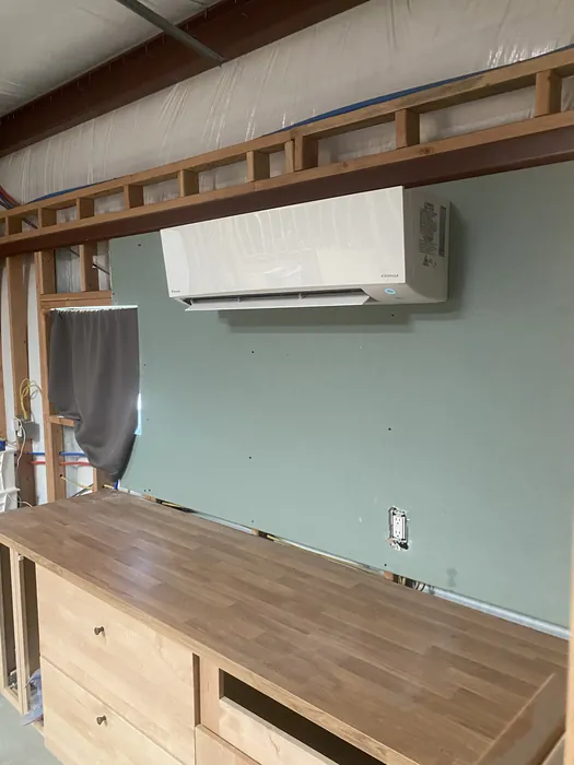 A white wall-mounted ductless mini-split indoor head installed high on a green drywall wall in a room under construction, with exposed wood framing on one side and finished wood flooring.
