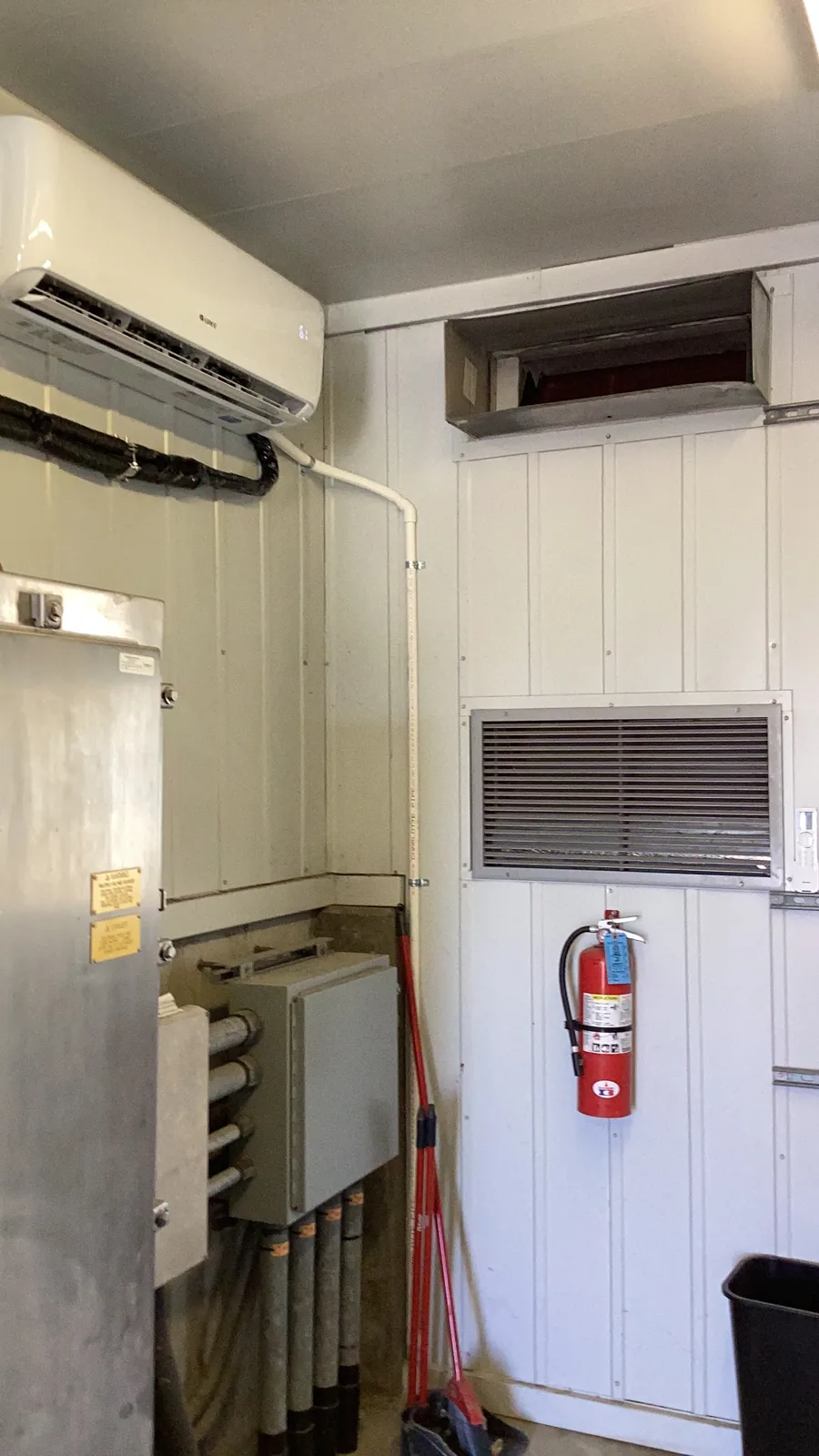 Indoor wall-mounted ductless mini-split air handler head beside a return grille and fire extinguisher in a High Desert mechanical room.