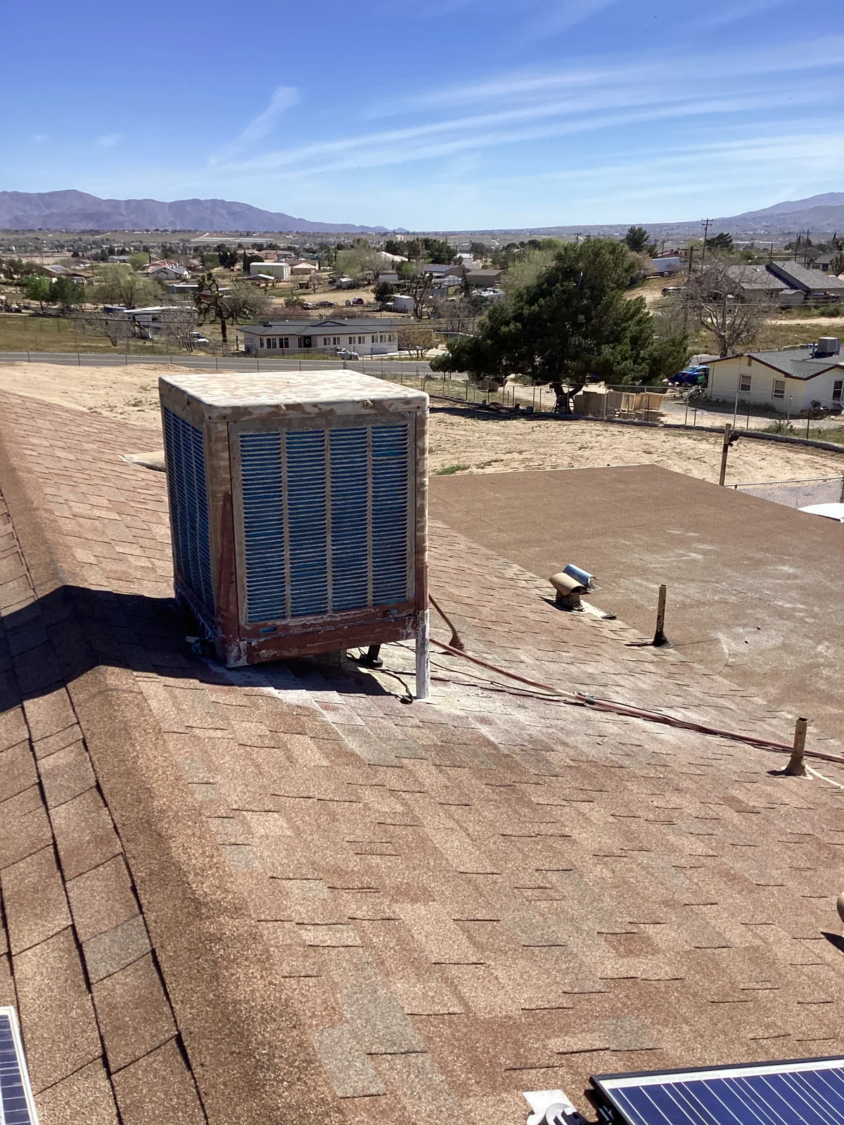 Rooftop evaporative swamp cooler mounted on a shingled roof with the High Desert valley and mountains in the background.