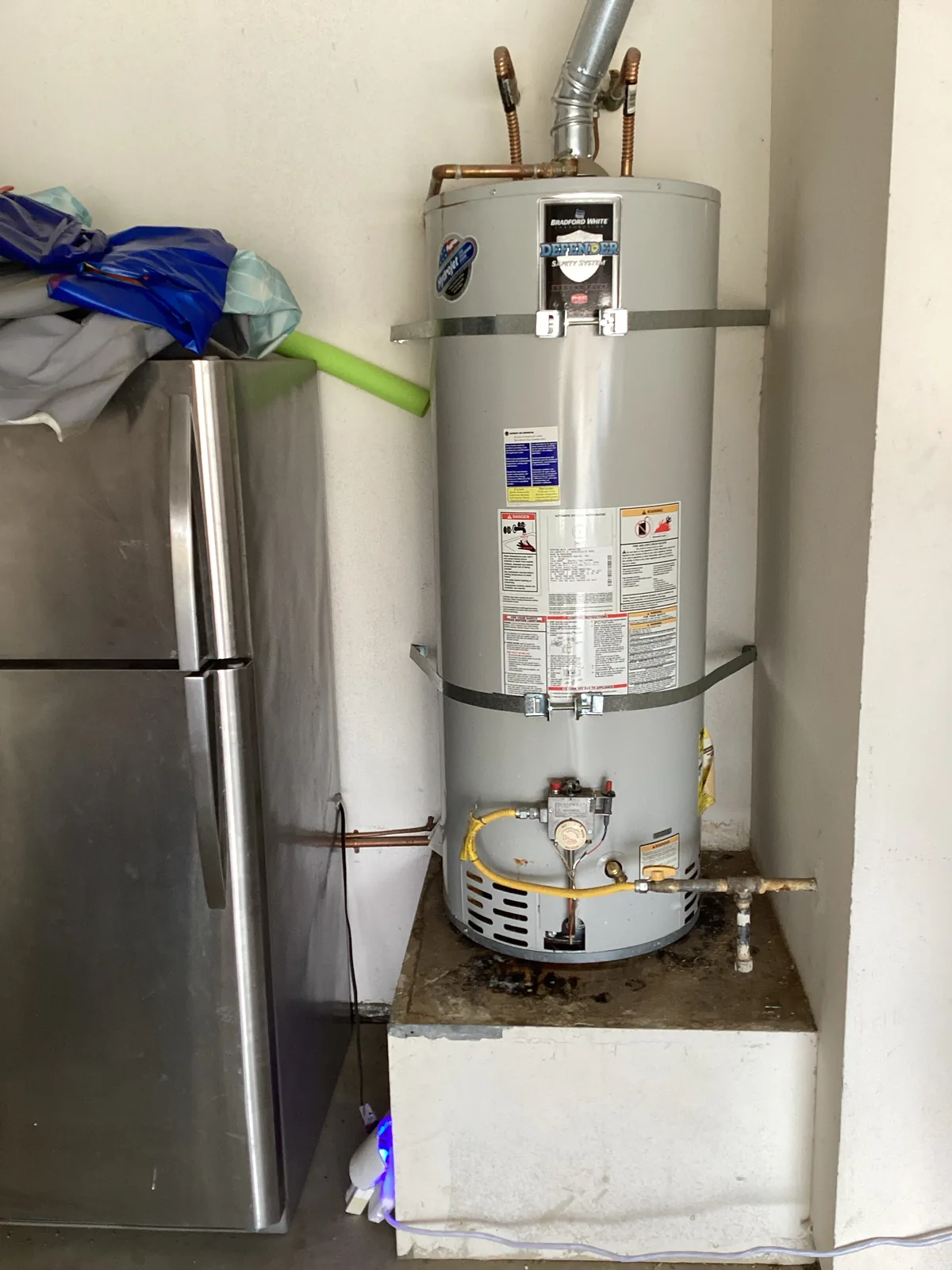 Bradford White Defender gas water heater seismically strapped on a raised platform beside a refrigerator in a garage.