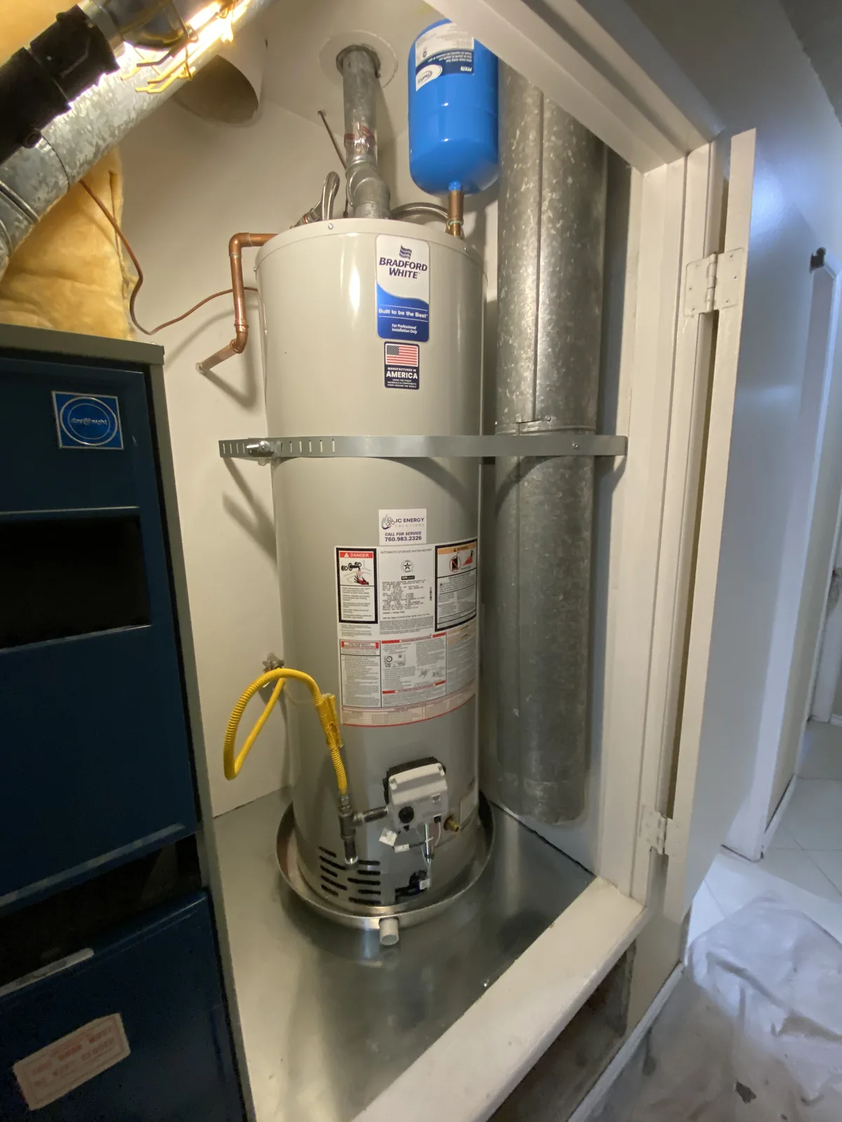 A Bradford White gas water heater tank with a blue expansion tank, copper piping, and a yellow gas flex connector installed in a closet above a blue furnace cabinet.